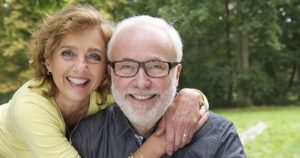 A husband and wife with implant retained dentures smiling with greenery behind them